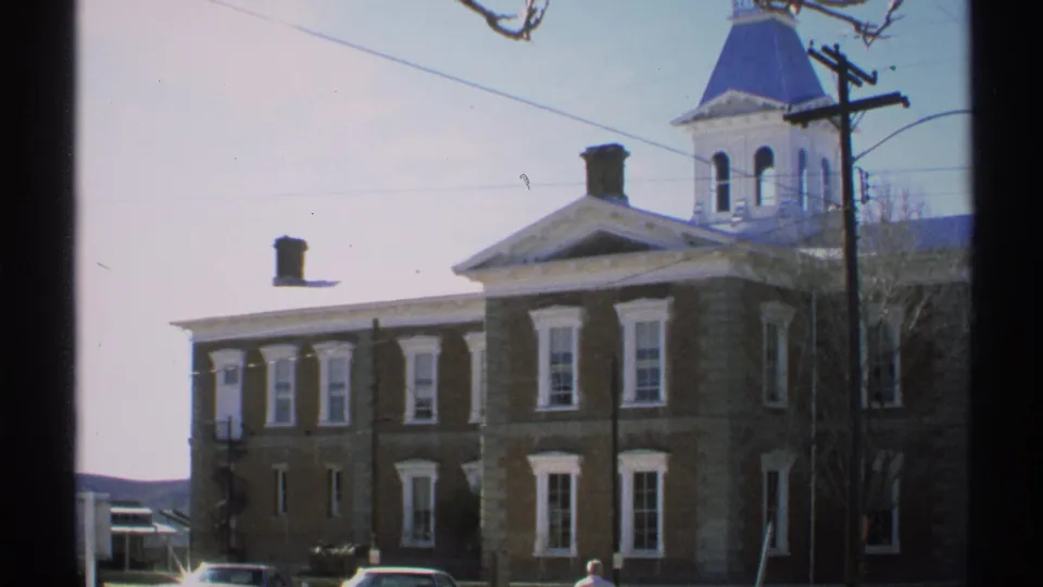 1972 Arizona home-movie view of Tombstone Courthouse, framed by a utility pole, overhead wires, bare tree branches, and parked cars below.