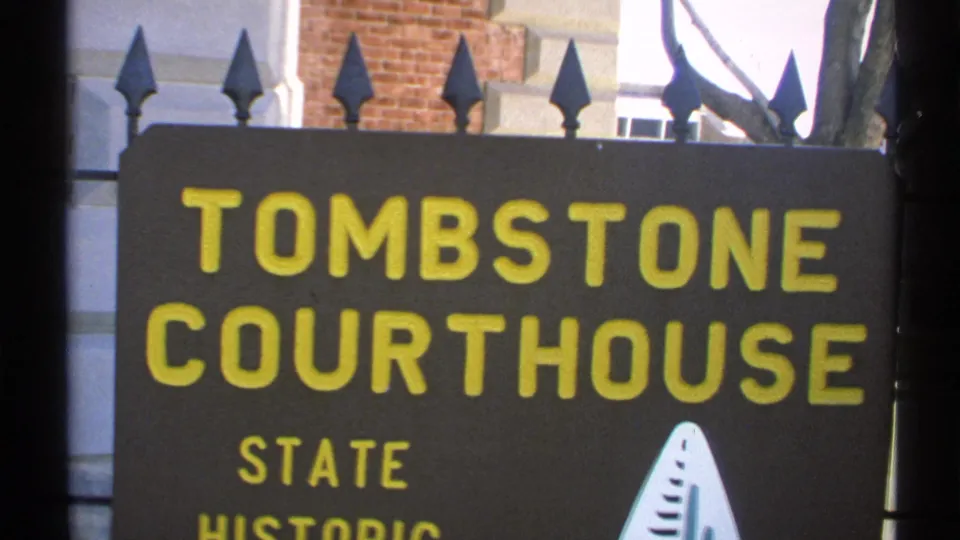 Close 1972 Arizona 8mm frame showing the Tombstone Courthouse state historic sign, iron fence points, brickwork, and part of a tree trunk.