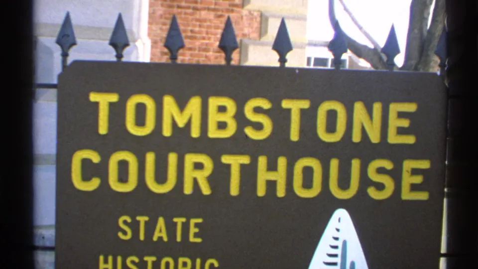 Close-up of the Tombstone Courthouse state historic sign in 1972 Arizona footage, with yellow lettering, fence points, brick, and white trim visible.
