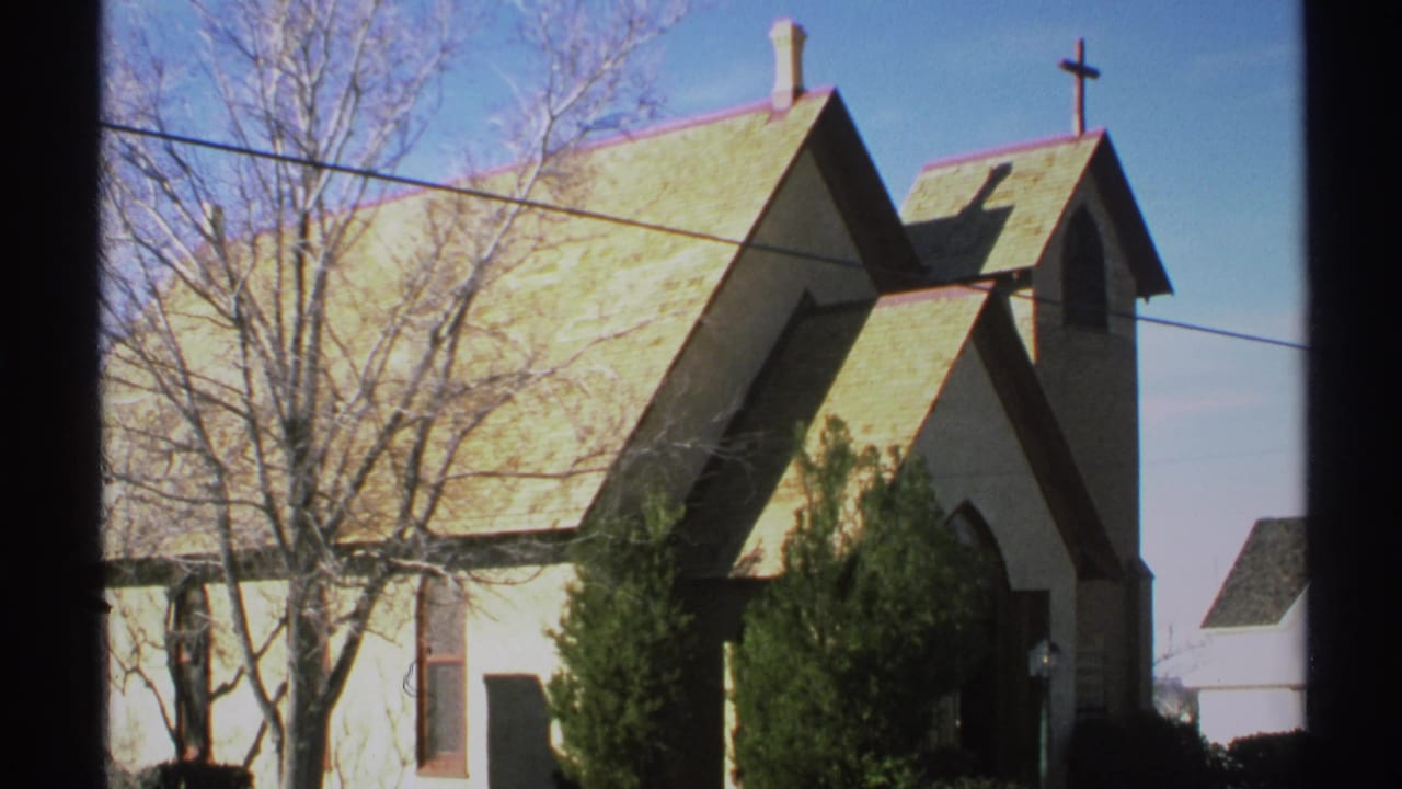 Small white church with green roof, cross, bare trees, shrubs, and power lines.