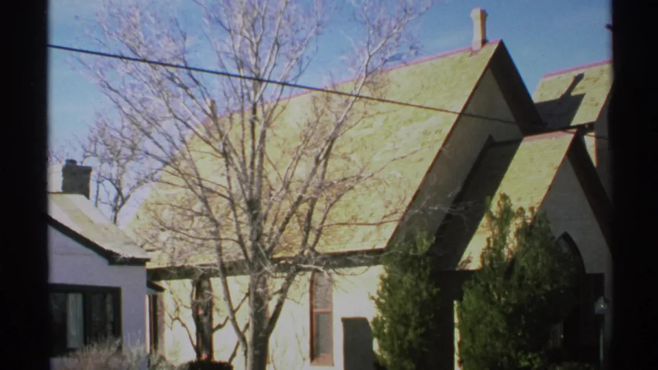 1972 Arizona 8mm frame centered on bare trees before a small white church with a green roof, shrubs, wires, and a nearby house edge.