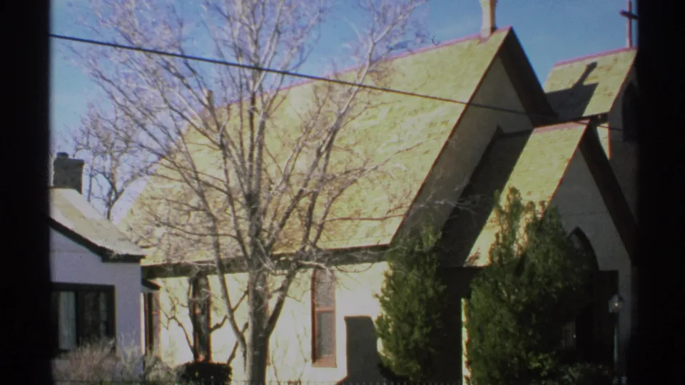 1972 Arizona 8mm frame showing the white church exterior, green roofline, bare winter trees, dense shrubs, overhead wires, and blue sky.