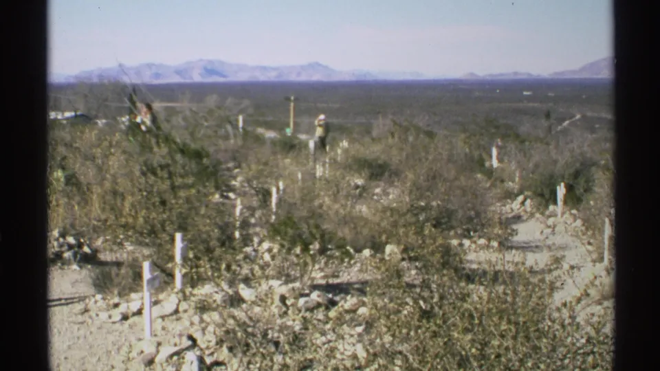 1972 Arizona 8mm frame of Boothill Graveyard, showing scrub-covered ground, white markers, distant visitors, and mountains.