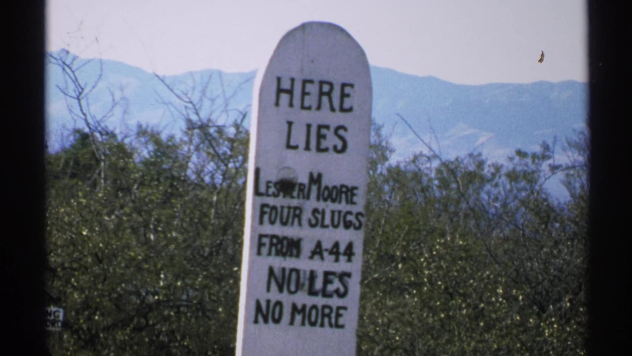 Close white tombstone with painted epitaph, desert brush, and mountains behind it.