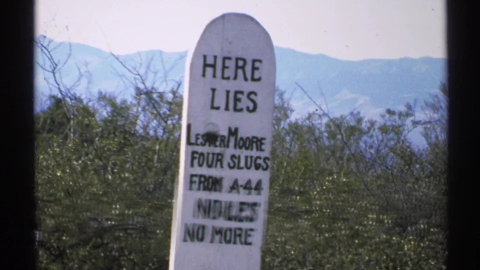 1972 Arizona 8mm frame of the tall white grave marker centered in the shot, with its epitaph partly blurred by motion and desert mountains behind it.