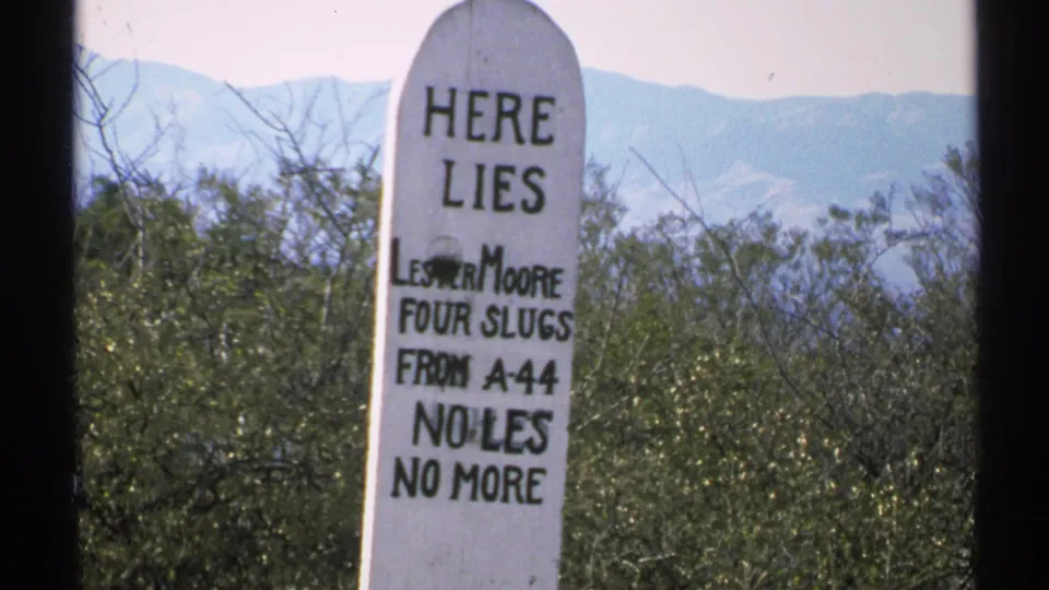 1972 Arizona 8mm frame showing the grave marker more clearly, including HERE LIES and lines of the Lester Moore epitaph against desert brush and mountains.