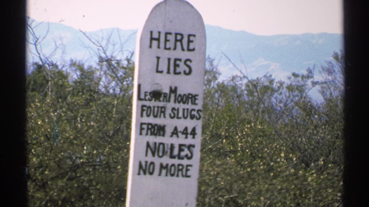 1972 Arizona 8mm frame of a white wooden marker reading Here Lies Lester Moore, set against shrubs and distant mountains.