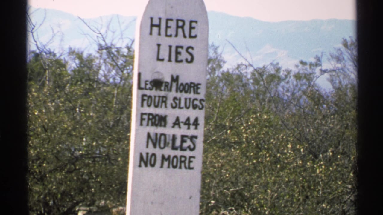 White gravestone-shaped sign reading Here Lies Lester Moore among desert shrubs.