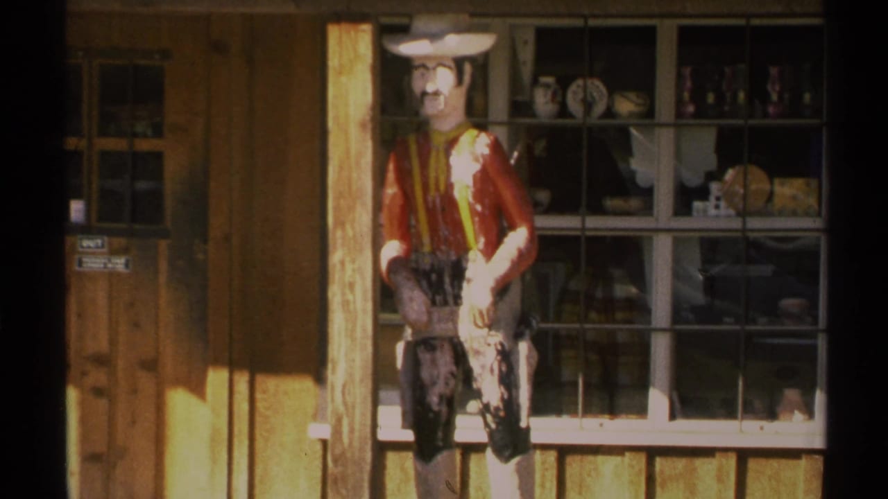 1972 Arizona 8mm frame showing a faded cowboy mannequin in a white hat beside a rustic storefront window with pottery and souvenirs.