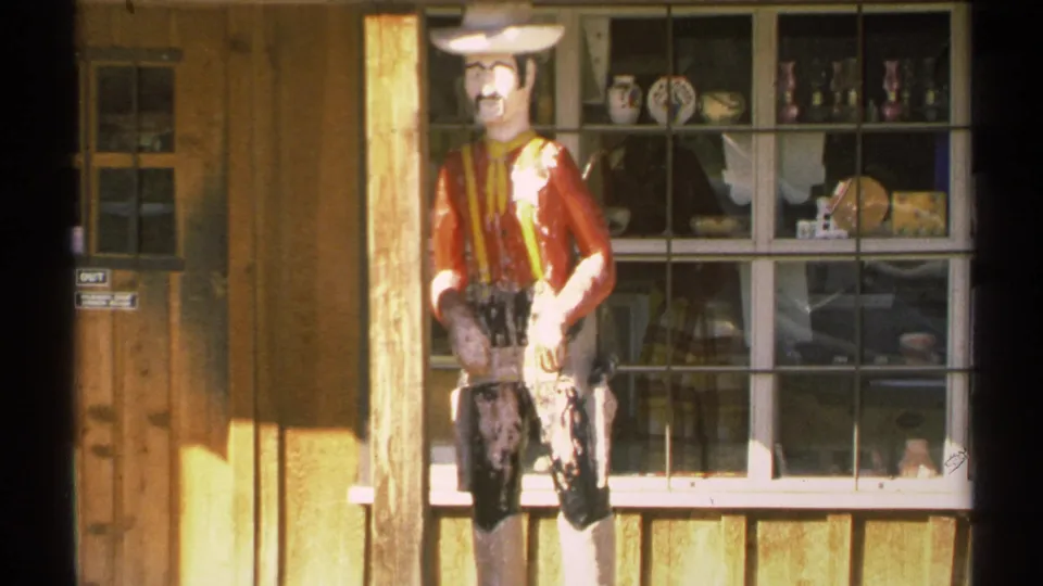 Closer 1972 Arizona 8mm view of the weathered cowboy figure, wood facade, door, and window display with pottery and small objects.