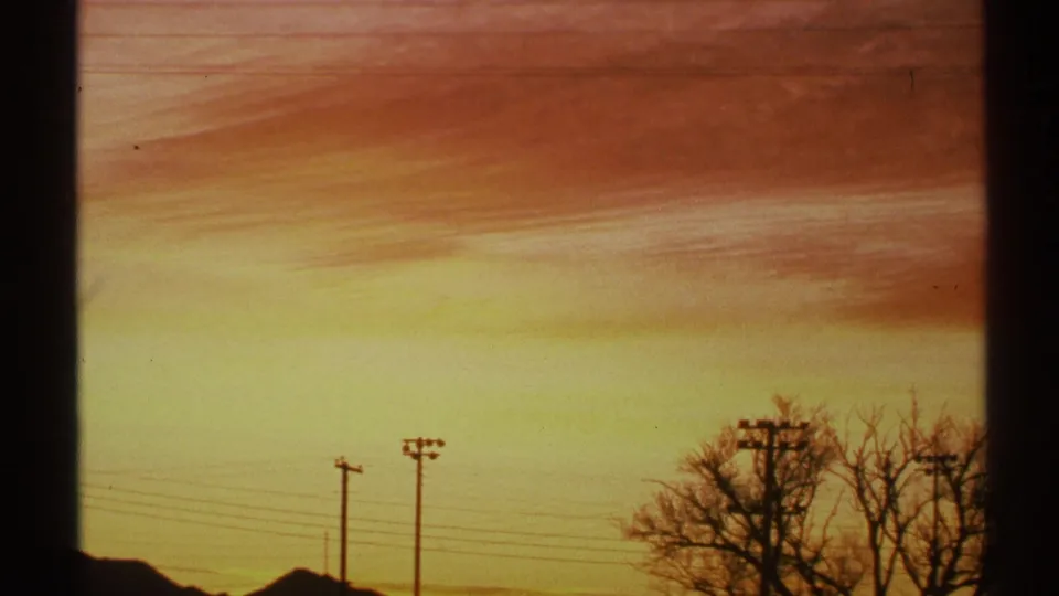 1972 Arizona 8mm frame of a warm sunset sky with thin clouds over dark utility poles, wires, bare trees, and low rooflines.