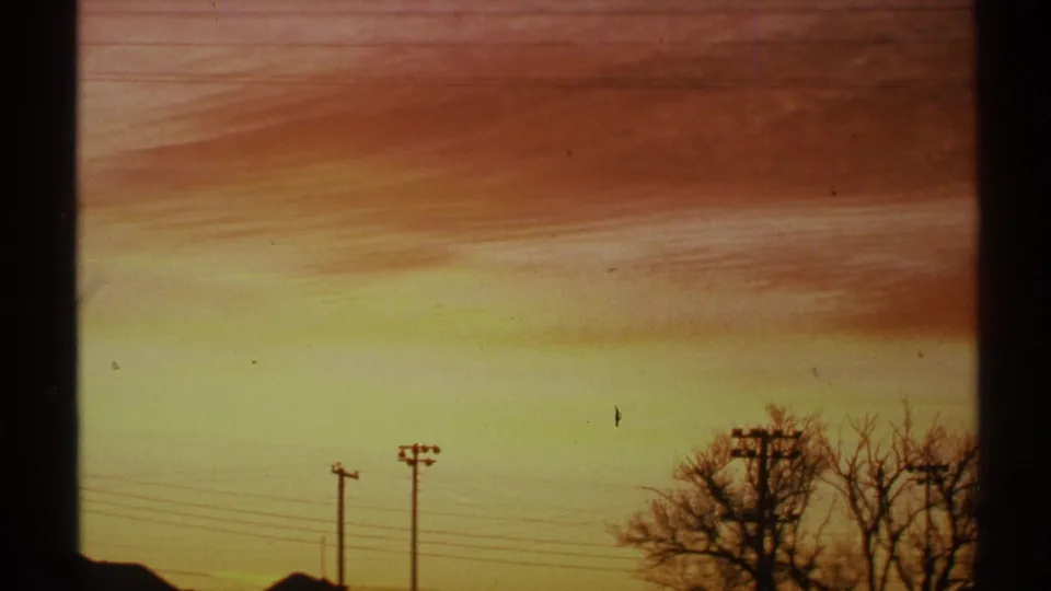 1972 Arizona 8mm view of a yellow-orange dusk horizon beneath thin clouds, with poles, wires, rooftops, and bare trees in silhouette.