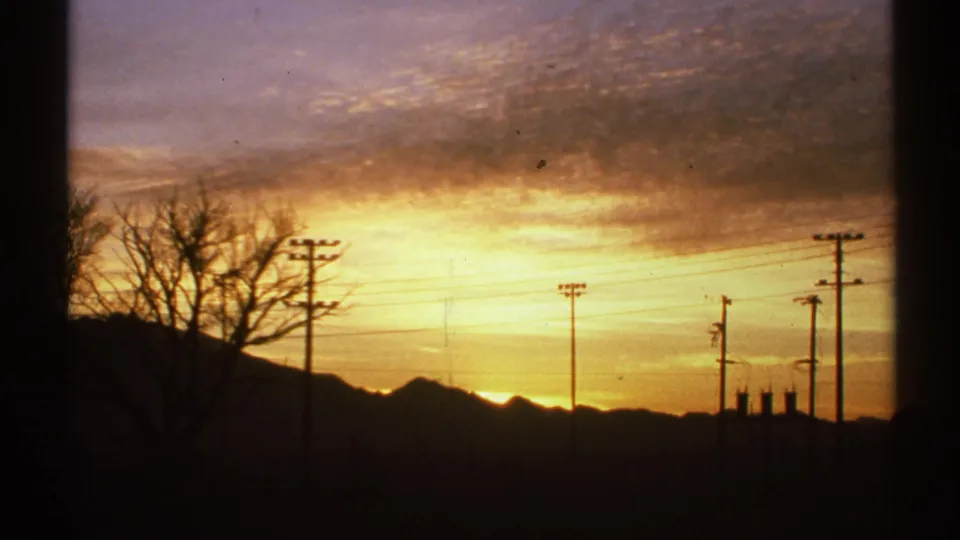 1972 Arizona 8mm frame showing utility poles, a bare tree, low structures, and a mountain horizon beneath sunset clouds.