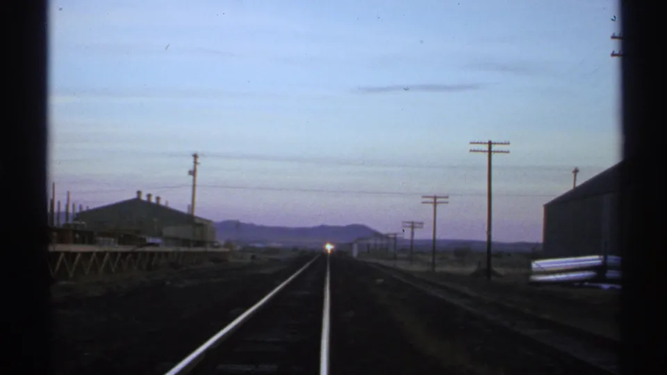 Forward view along Arizona railroad tracks at twilight, with low rail-side buildings, utility poles, and a distant train headlight.