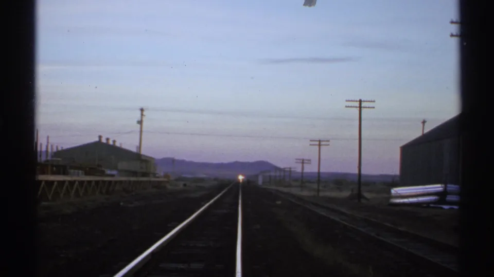 Twilight railroad view in Arizona, showing tracks, utility poles, low structures, mountains, and a small distant train light.