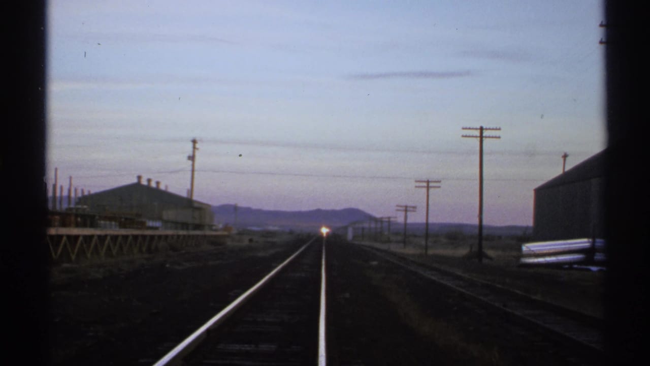Railroad tracks recede toward a distant headlight at dusk, framed by dark vertical edges.