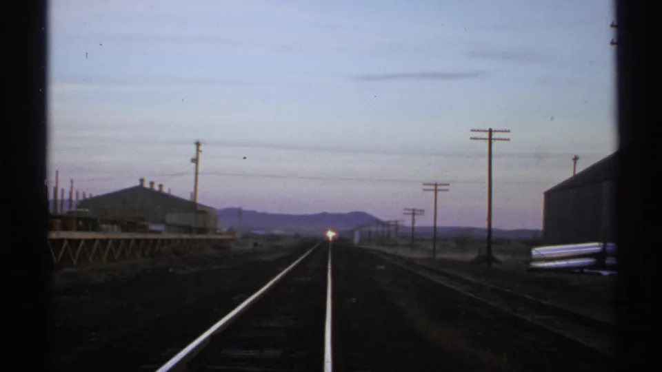 1972 Arizona 8mm frame showing rails, utility poles, trackside buildings, distant mountains, and a small train headlight at dusk.
