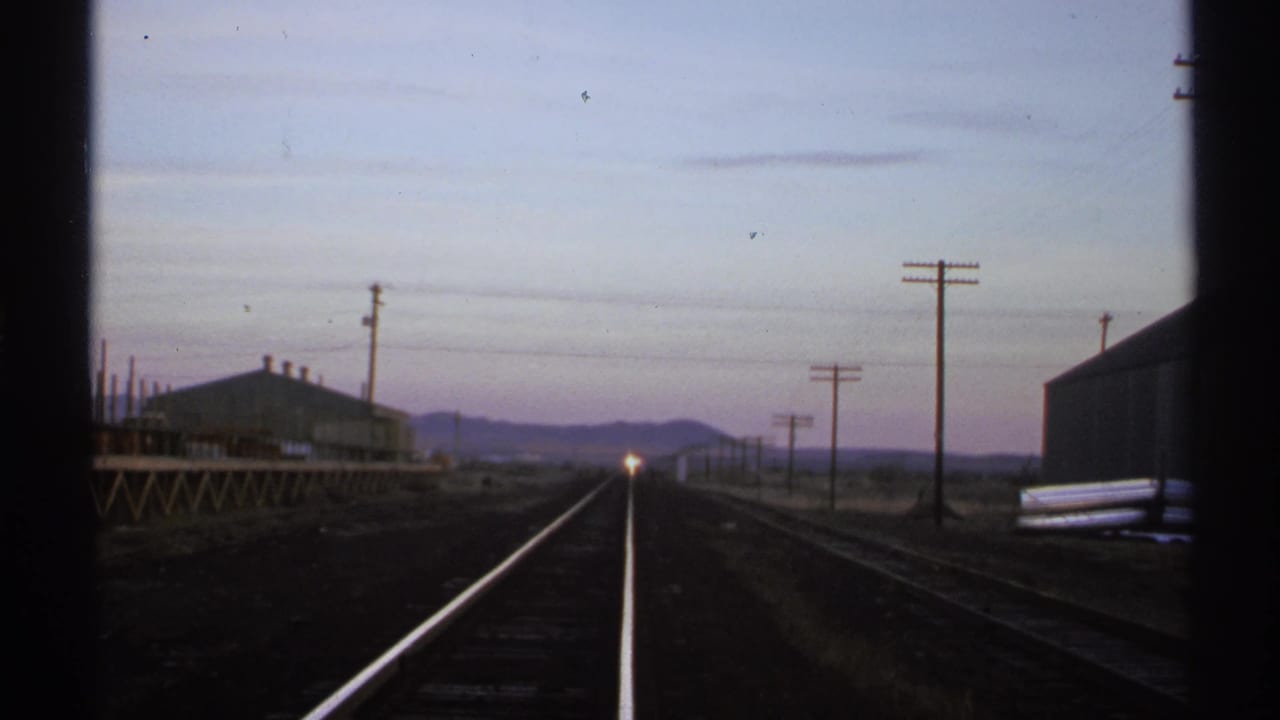 A small train headlight glows on the horizon between rails and trackside buildings.