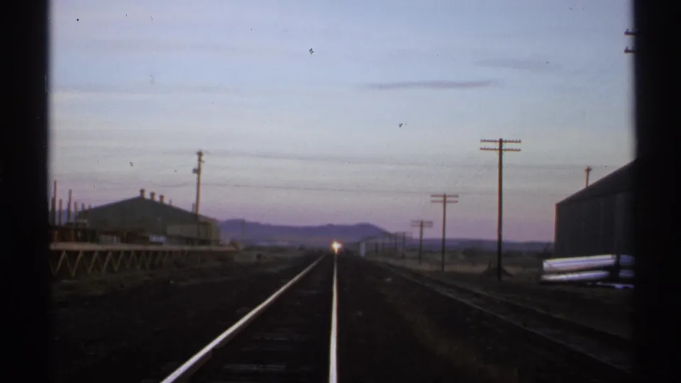 1972 Arizona rail scene with a freight train still distant, its headlight centered near the tracks as utility poles line the route.