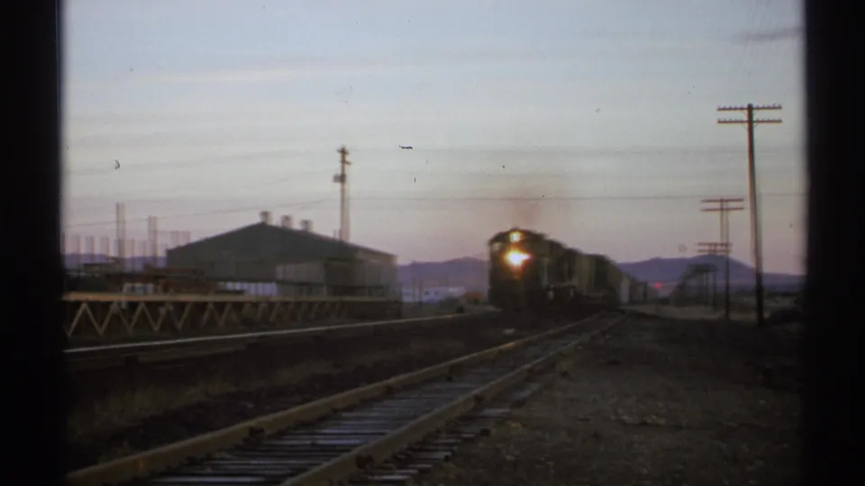 1972 Arizona 8mm frame showing a freight locomotive nearing the station area, with rails, sheds, poles, and mountains behind it.