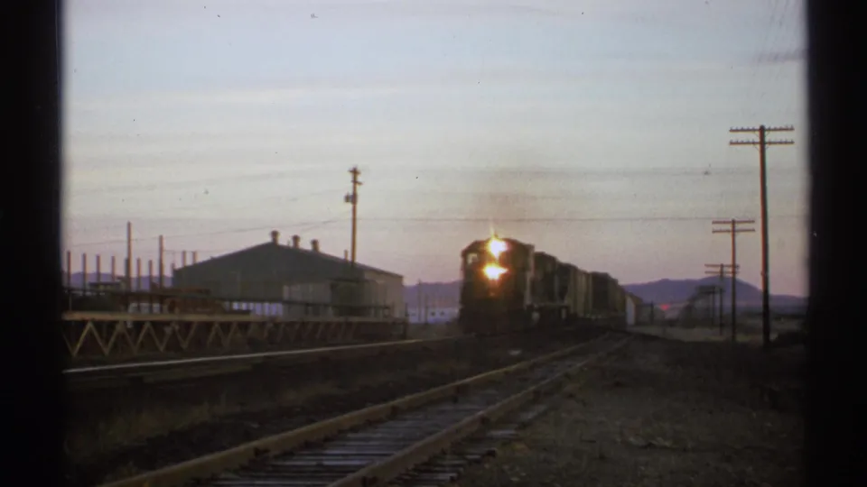 1972 Arizona 8mm frame of a freight locomotive arriving near the camera, with trackside structures and utility poles in low evening light.