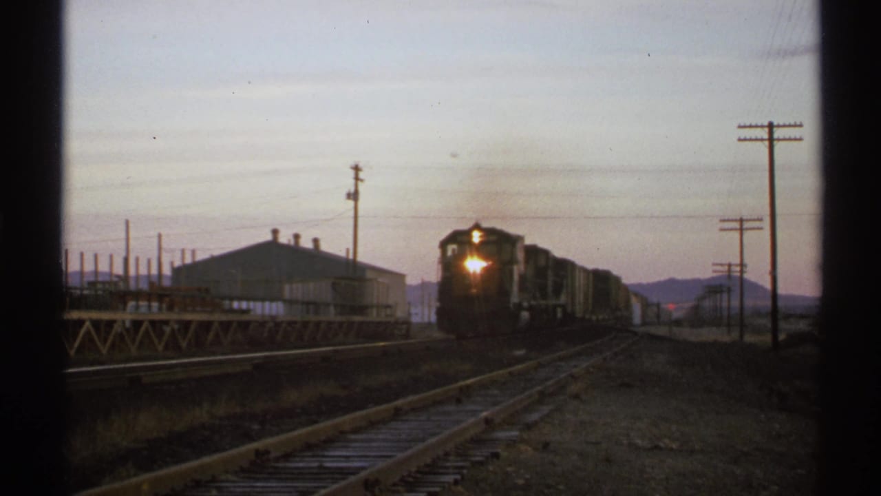 1972 Arizona 8mm frame of a freight train approaching at dusk, with rails, utility poles, an industrial building, and distant mountains.