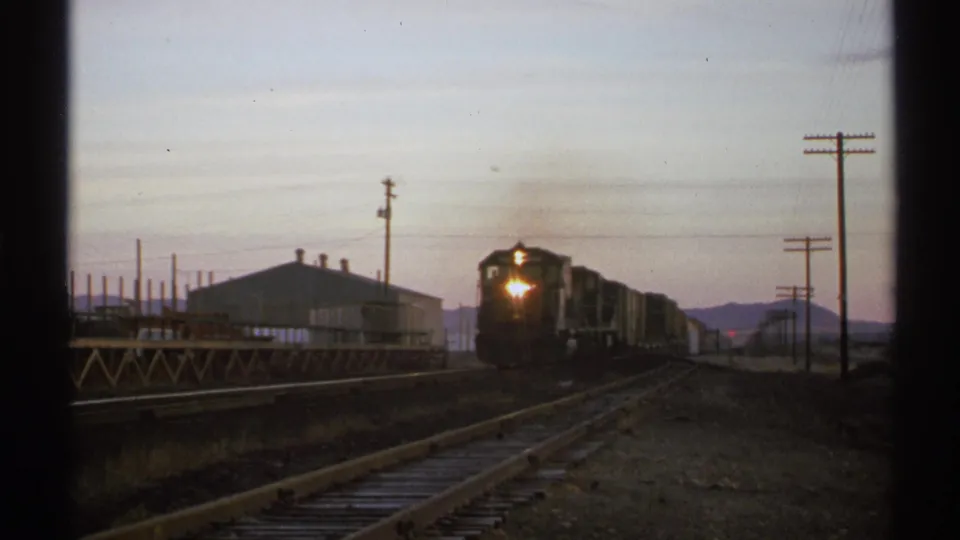 1972 Arizona 8mm frame of a freight train approaching at dusk, with rails, utility poles, an industrial building, and distant mountains.
