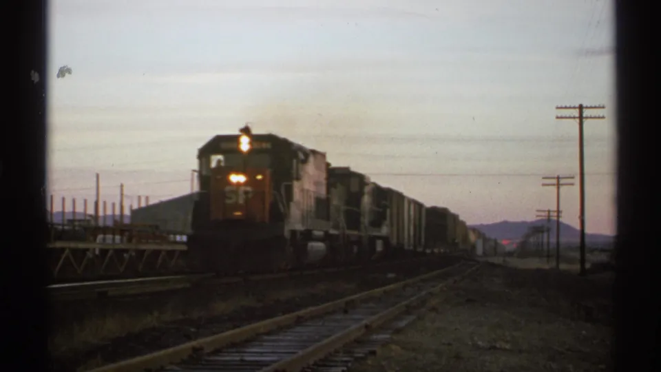 1972 Arizona 8mm frame of a close freight locomotive with visible SP lettering, followed by freight cars beside rails and utility poles.