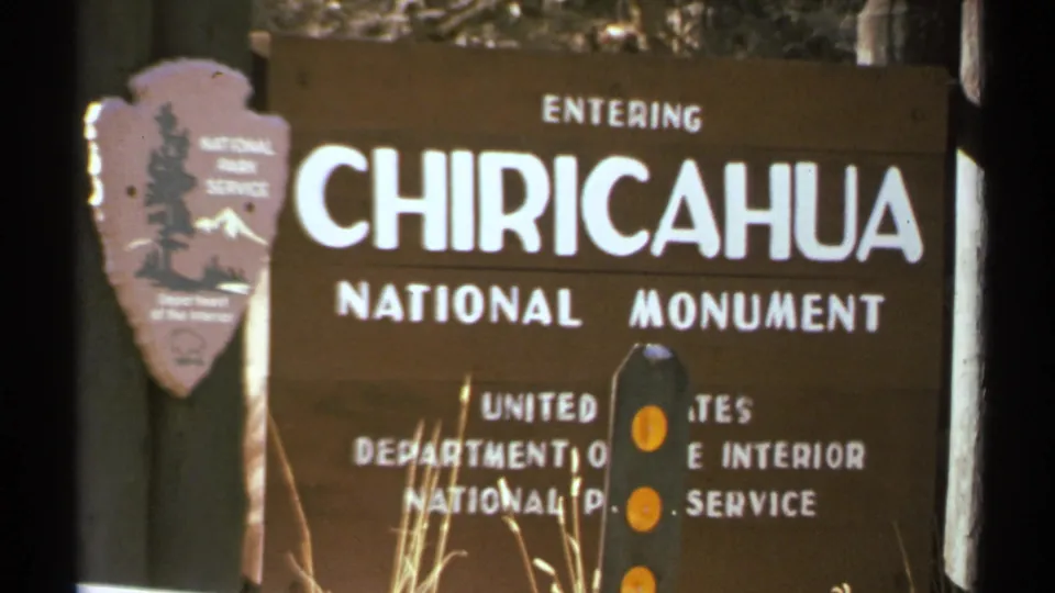 1972 Arizona frame holding on the Chiricahua National Monument entrance sign with posts, dry grass, and trees around it.