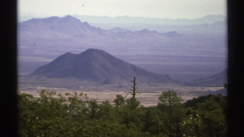 1972 Arizona home-movie frame of a broad desert valley, rounded hill, foreground trees, and hazy distant ranges.