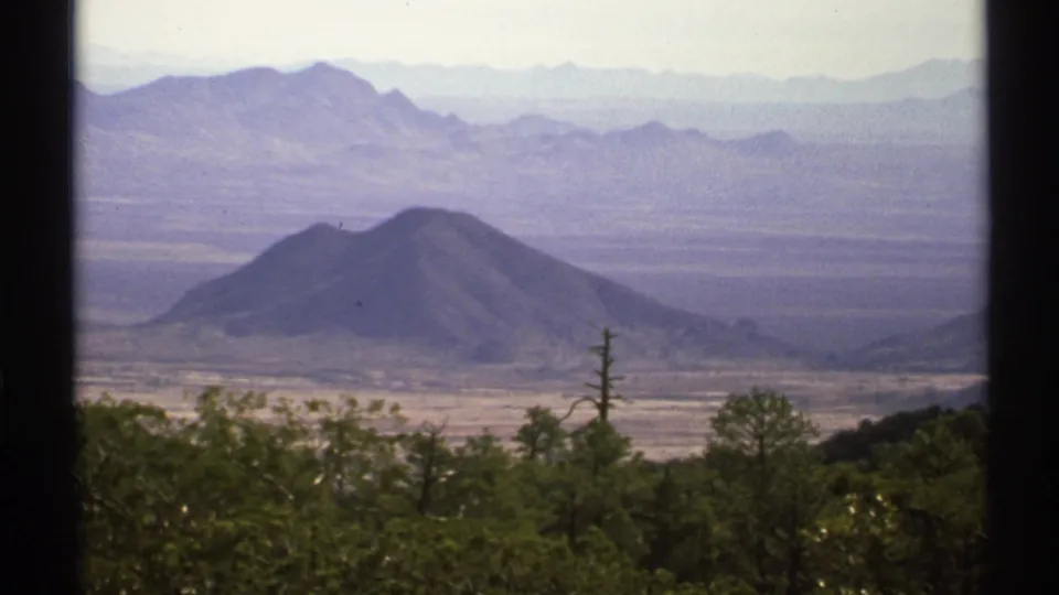 1972 8mm frame of an Arizona desert vista with a dark conical hill, layered ranges, scrubby foreground trees and hazy sky.