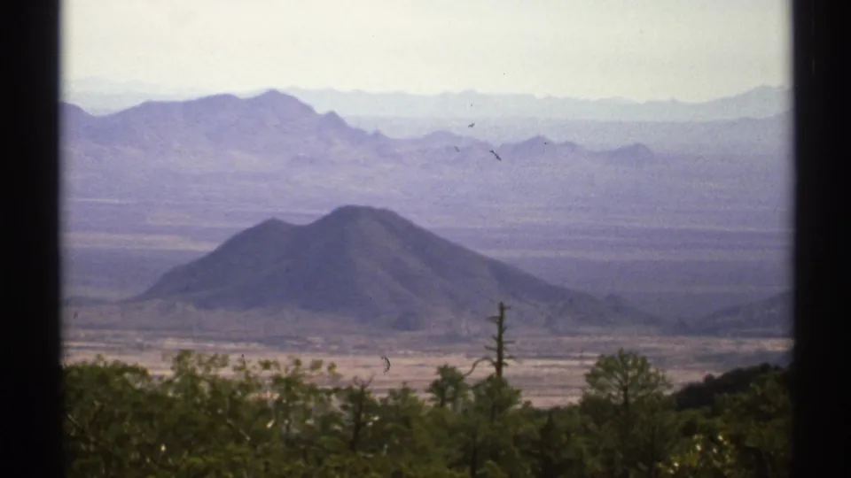 1972 8mm view across Arizona desert terrain toward a conical hill backed by hazy layered mountain ranges.