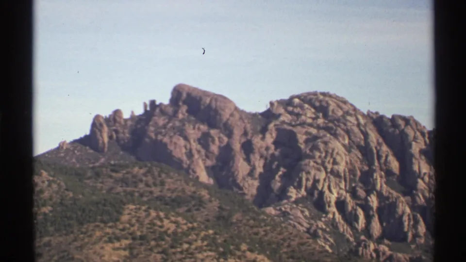1972 8mm frame showing jagged Arizona rock formations rising above scrub-covered slopes beneath a pale blue sky.