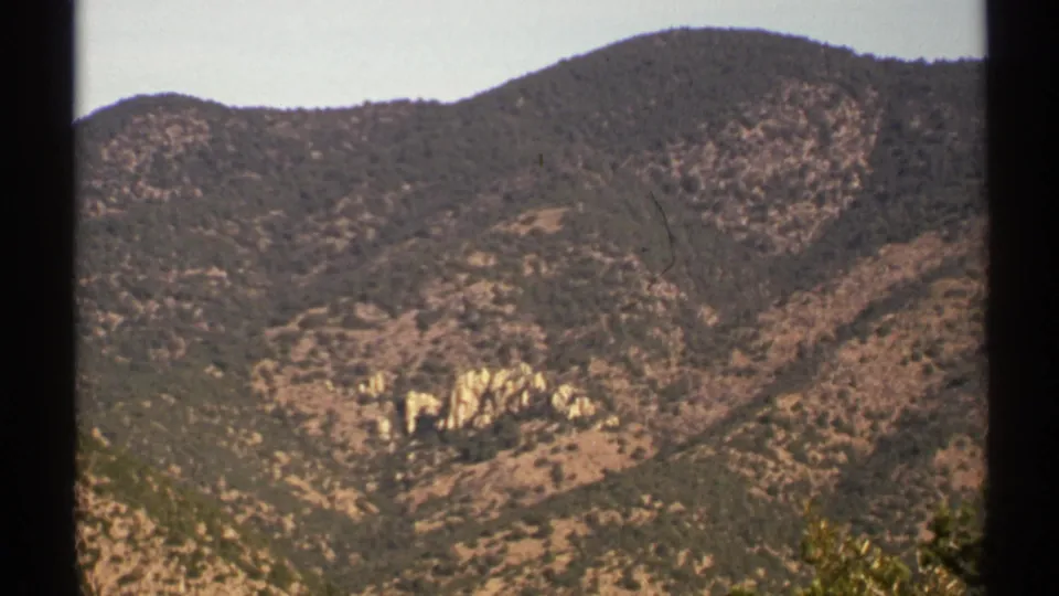 1972 Arizona 8mm frame showing rounded desert hills, scrub vegetation, and pale rock outcrops below a darker ridge.