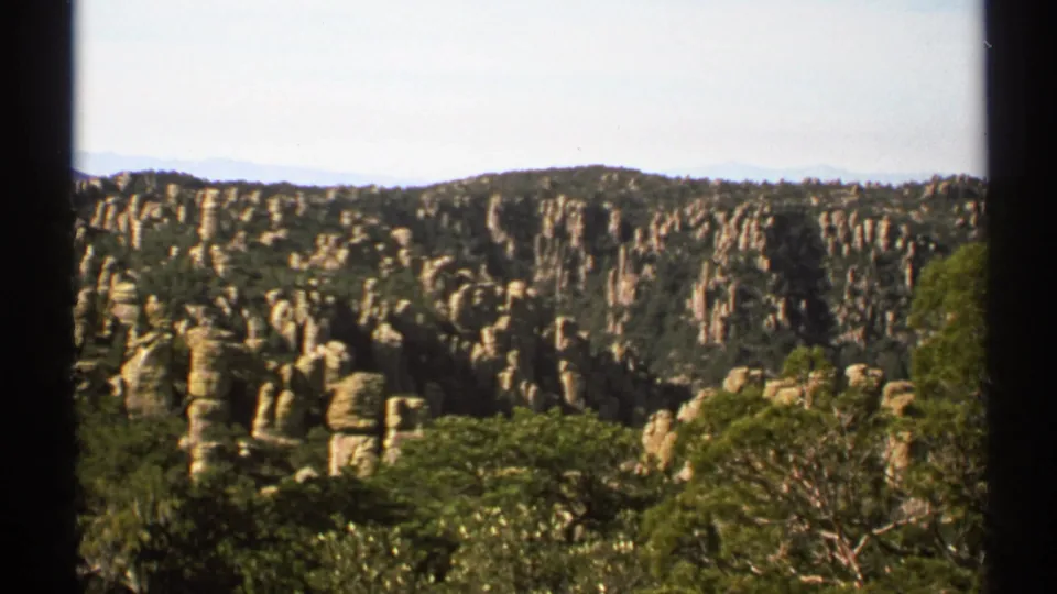 1972 Arizona 8mm frame of vertical rock formations rising above dense green trees in a canyon landscape.