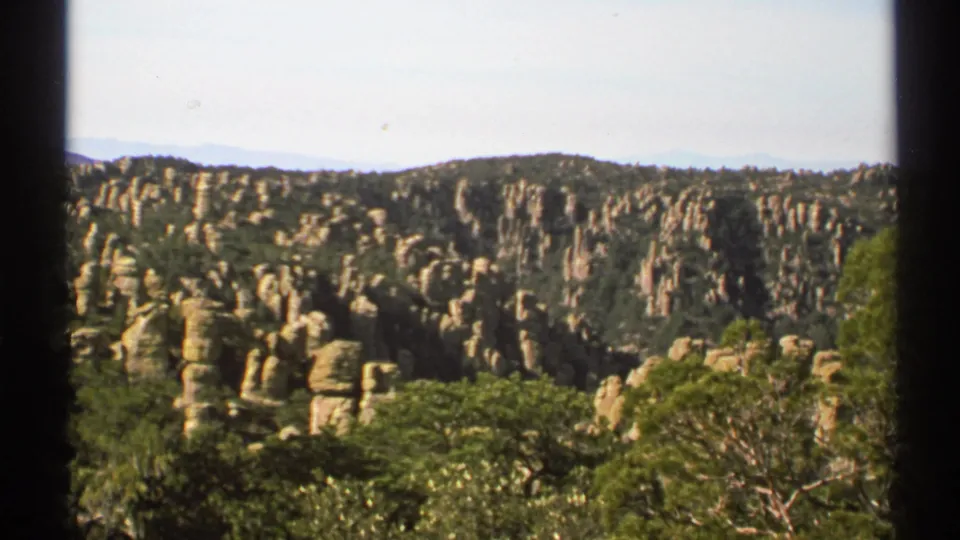 Wide 1972 home-movie view of Arizona stone pillars among green trees, framed by dark 8mm film edges.