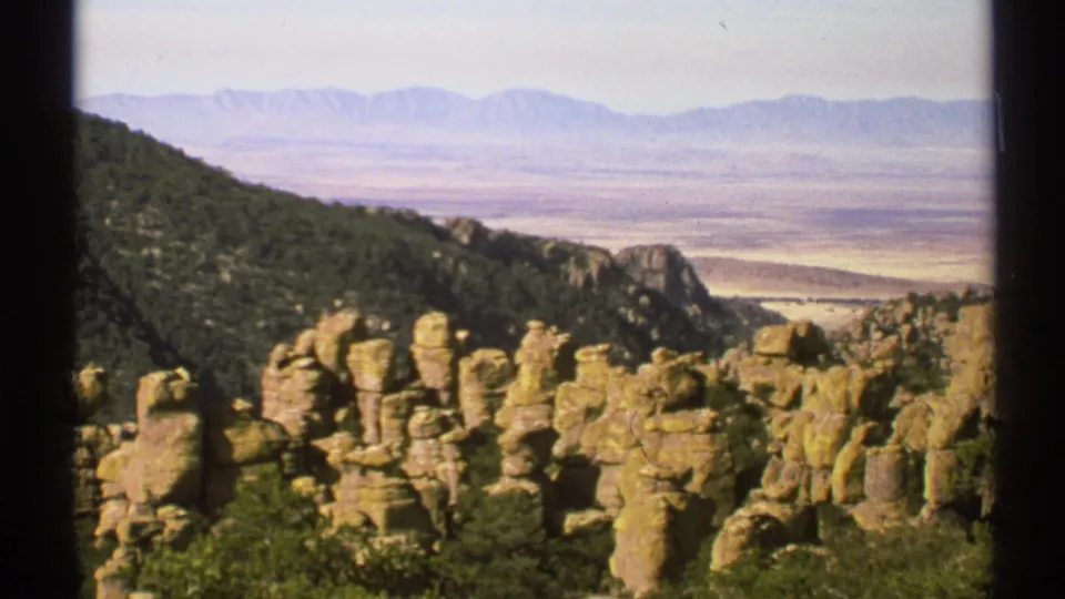 1972 Arizona landscape view from an overlook, with rock formations, a wooded hillside, broad valley, and distant mountains.