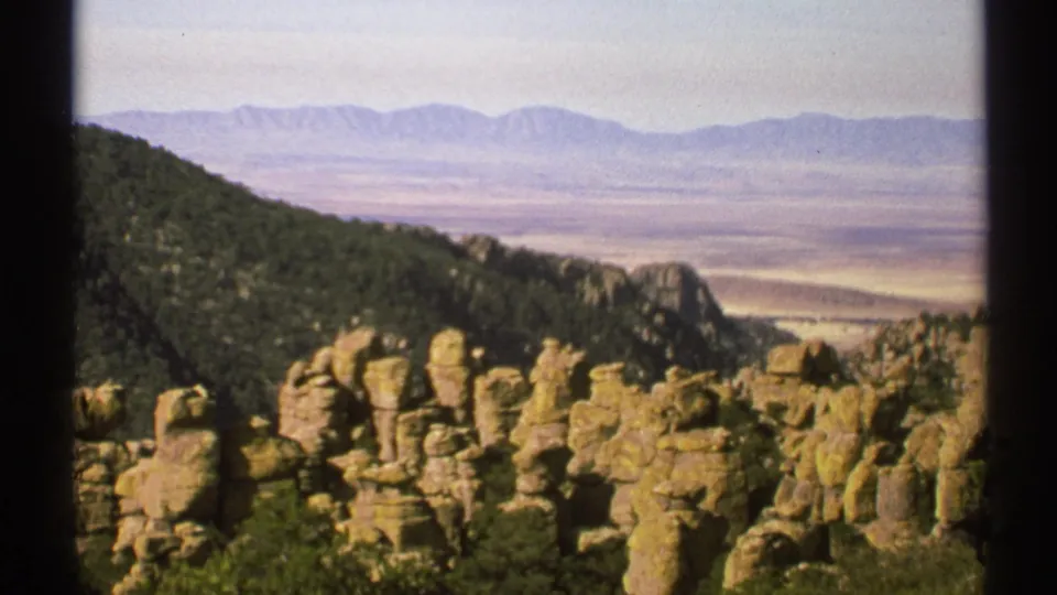 Arizona 8mm frame showing foreground rock formations and brush with a hazy valley and distant mountain ranges beyond.