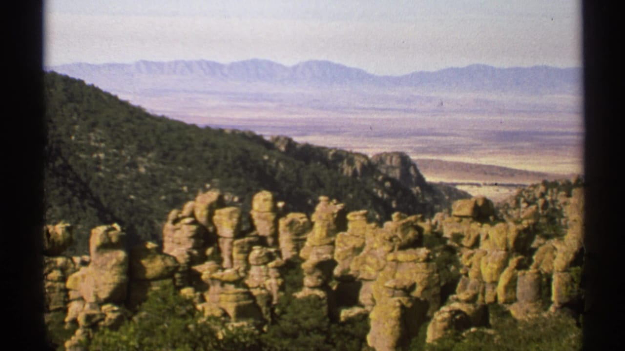 1972 Arizona 8mm frame showing tan rock formations, green hillside, pale desert valley, and distant mountains under a hazy sky.