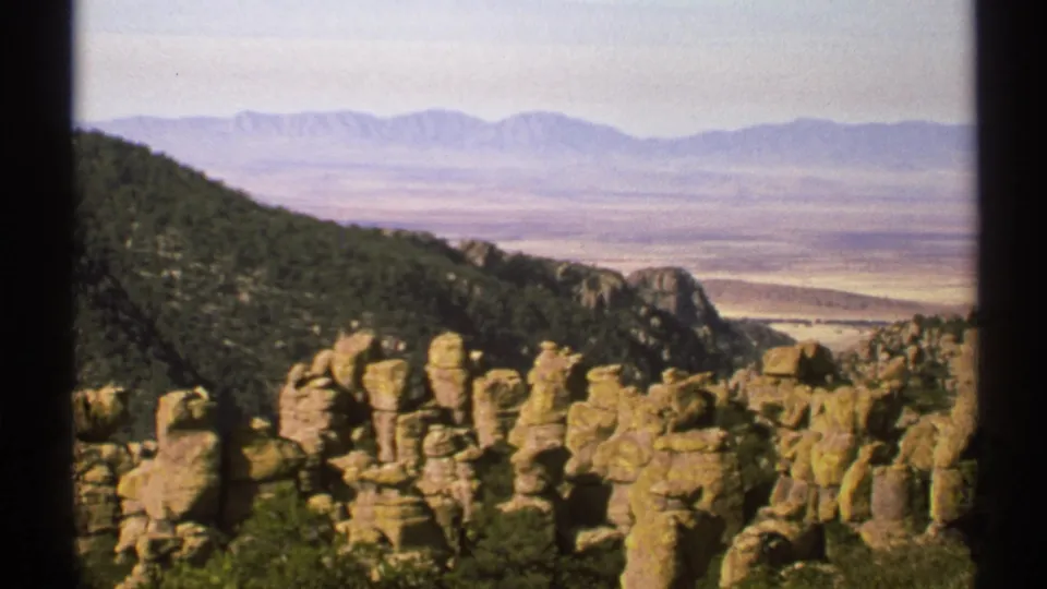 1972 Arizona 8mm frame with rugged rock spires in the foreground, a wooded slope, open desert valley, and blue-gray mountains.