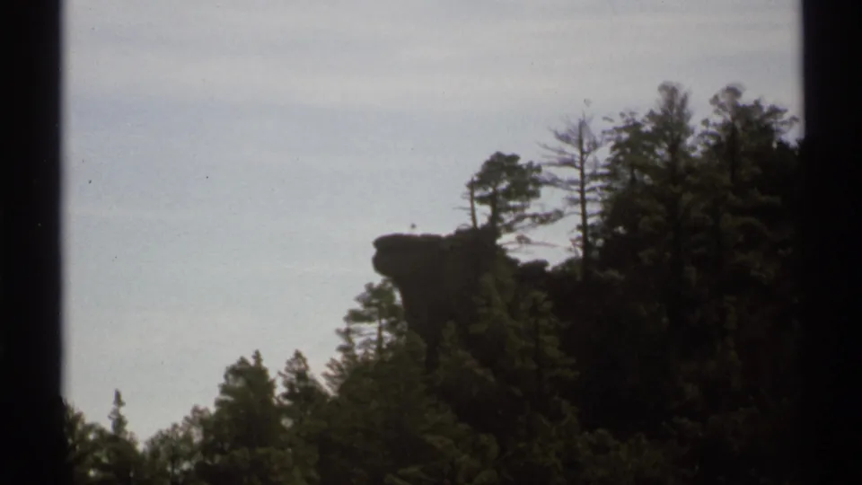 1972 Arizona home-movie frame with a dark gate edge, wooded slope, and protruding rock outcrop under a washed pale sky.