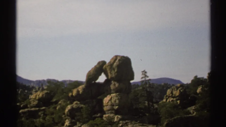 1972 Arizona 8mm frame showing a natural rock arch, rugged boulders, sparse vegetation, and distant low hills.