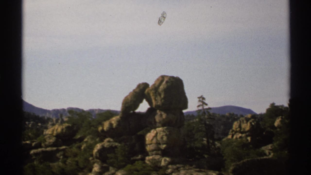 1972 Arizona 8mm frame showing a weathered rock arch among boulders, scrub vegetation, distant mountains, and pale sky.