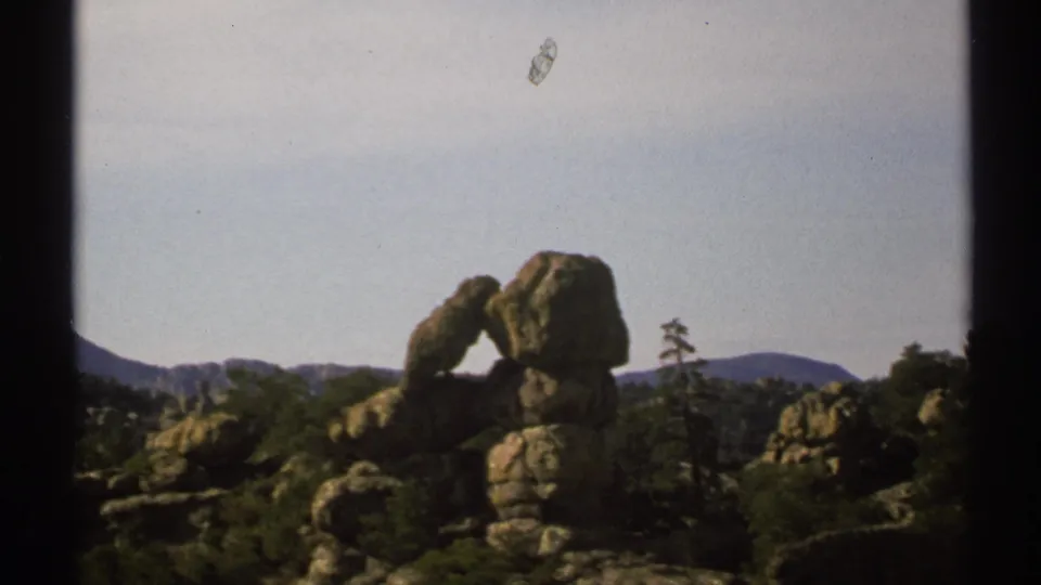 1972 Arizona 8mm frame showing a weathered rock arch among boulders, scrub vegetation, distant mountains, and pale sky.