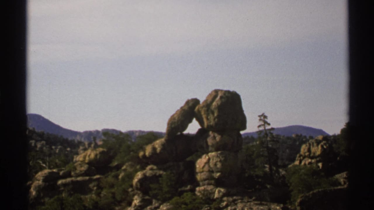 Balanced rock arch framed by shrubs, boulders, and low mountains under a pale sky.