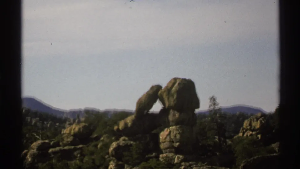 Arizona 1972 8mm frame with the rock arch slightly lower in view, surrounded by brush, boulders, distant hills, and dark gate edges.