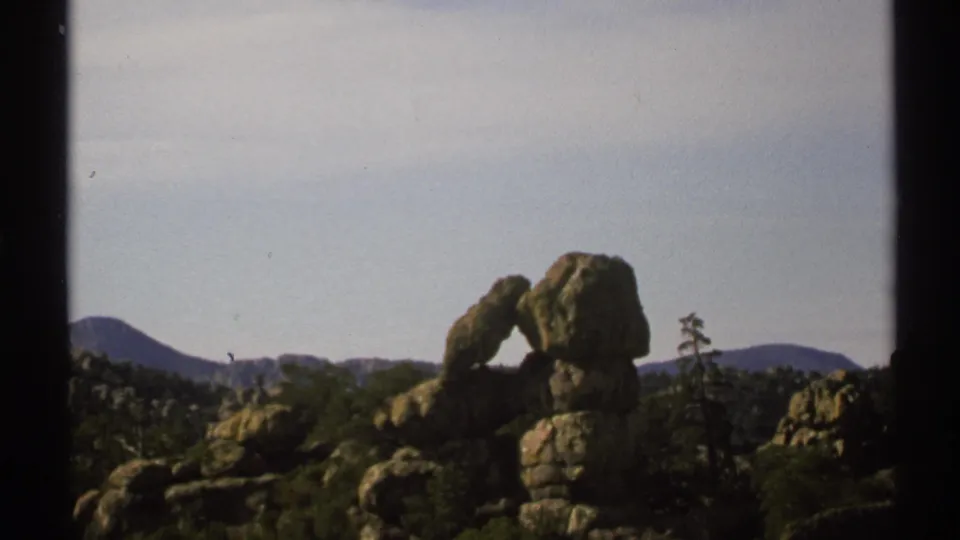 1972 Arizona 8mm still showing the rock arch and boulders with scrub vegetation, distant mountains, and a small bird-like speck in the sky.