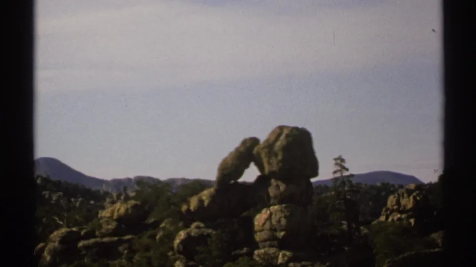 1972 Arizona 8mm frame showing a balanced-looking rock formation, wooded terrain, distant mountains, and pale sky.