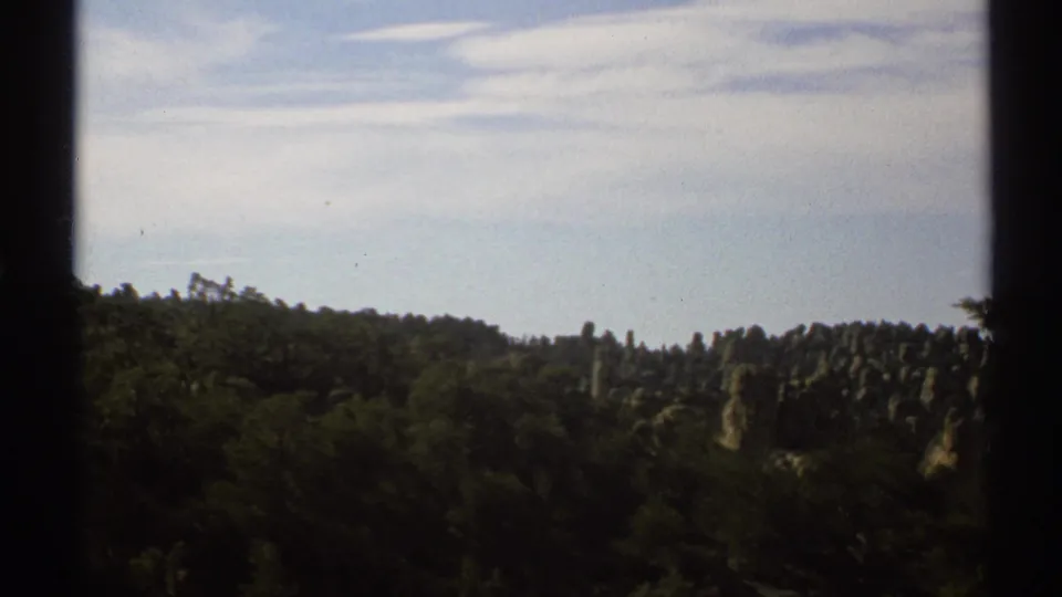1972 Arizona home-movie frame looking across wooded rocky terrain and low ridges under a pale blue sky.