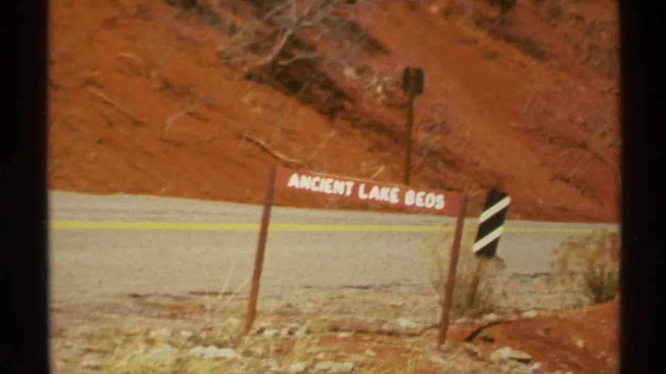 1972 Arizona roadside view with an Ancient Lake Beds sign, red earth hillside, road edge, dry brush, and marker post.